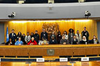 Malgosia Fitzmaurice and Andrew Pitts with Queen Mary LLM students on the stage of the the International Maritime Organization's assembly room. Behind them is the IMO logo on a wood paneled wall.