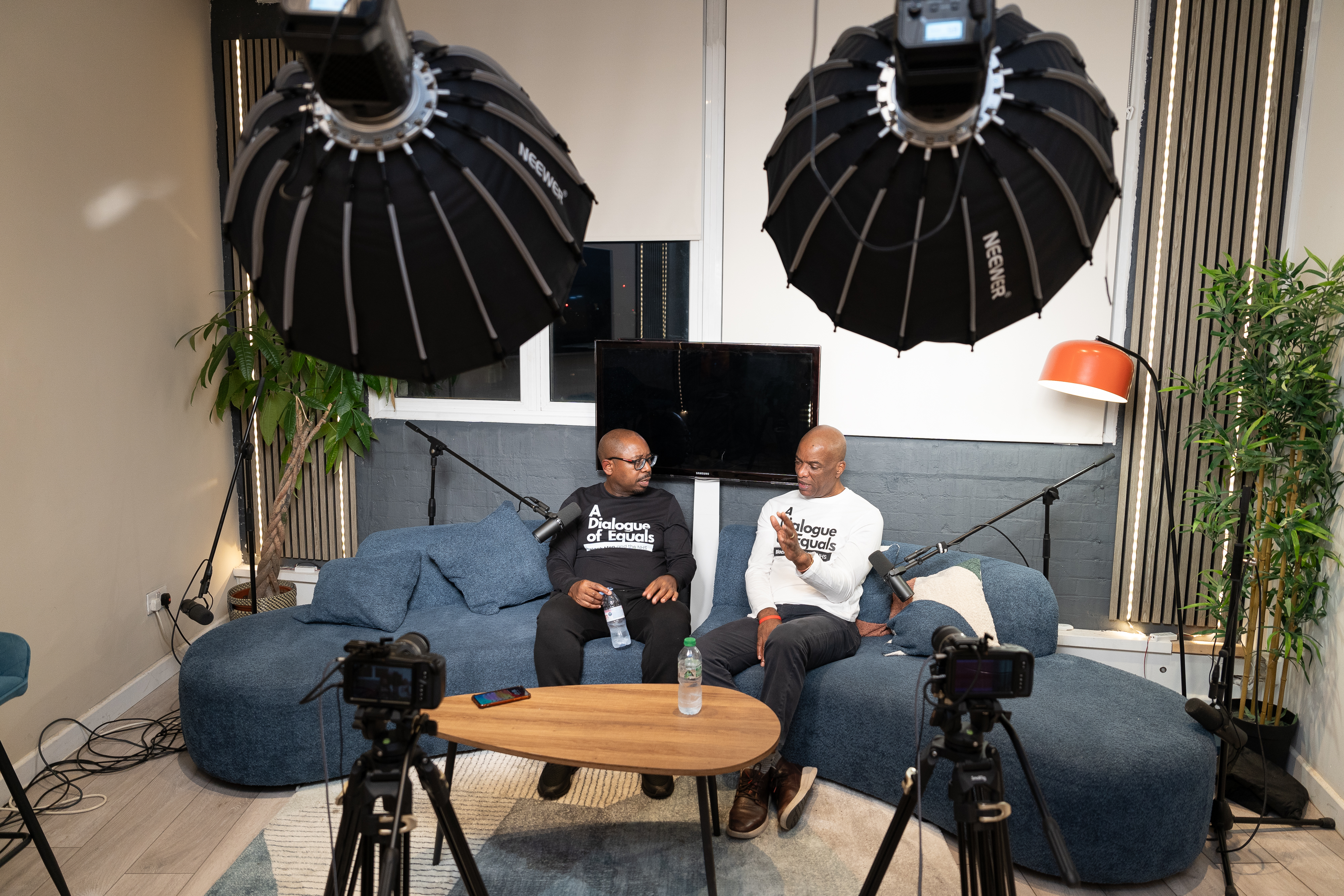 Two Black men sit on a sofa recording a podcast