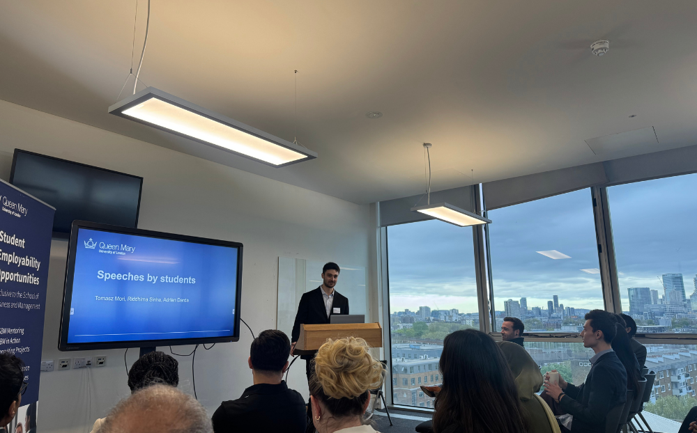 Image of a person speaking infront of a presentation in a room full of people sitting on chairs and the view of the london skyline in the background
