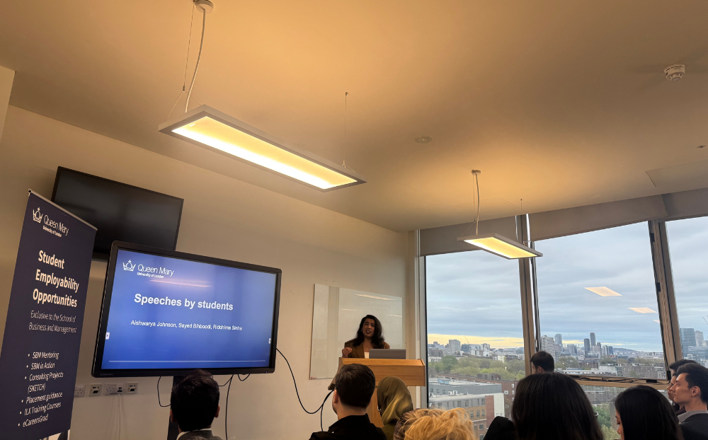 Image of a person speaking infront of a presentation in a room full of people sitting on chairs and the view of the london skyline in the background