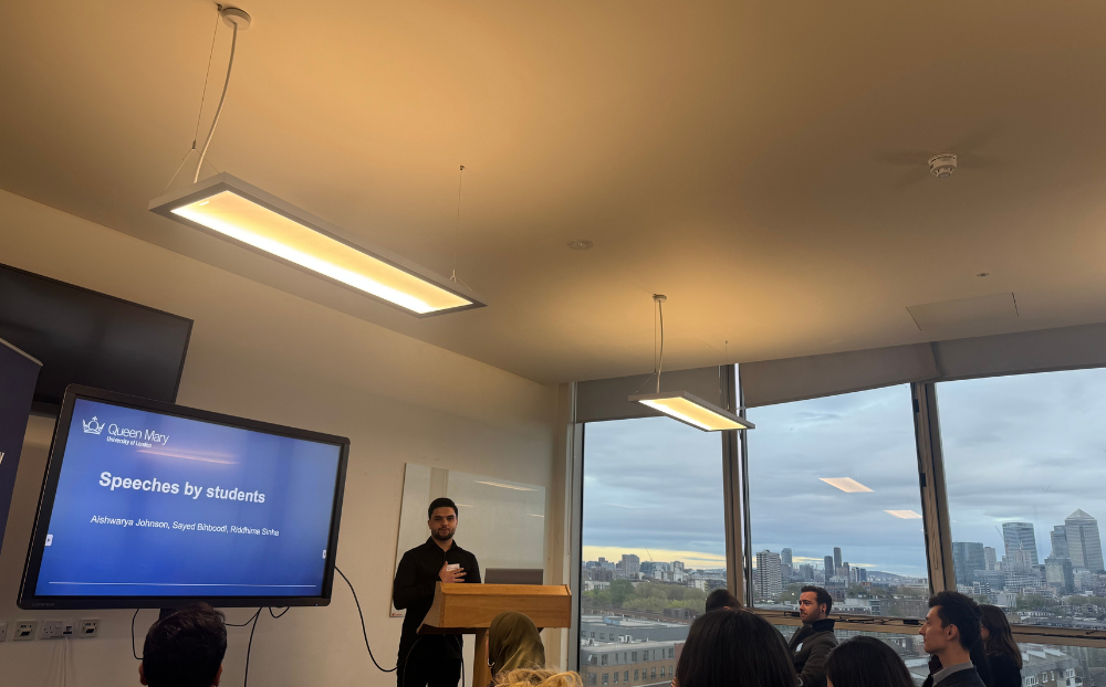 Image of a person speaking infront of a presentation in a room full of people sitting on chairs and the view of the london skyline in the background