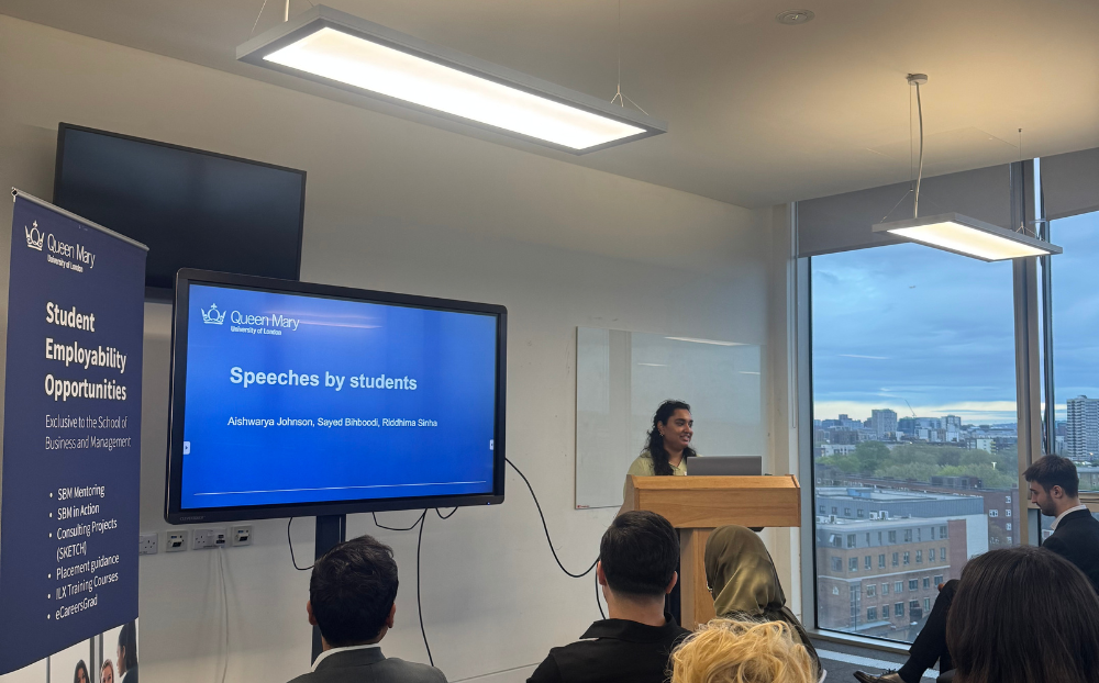 Image of a person speaking infront of a presentation in a room full of people sitting on chairs and the view of the london skyline in the background