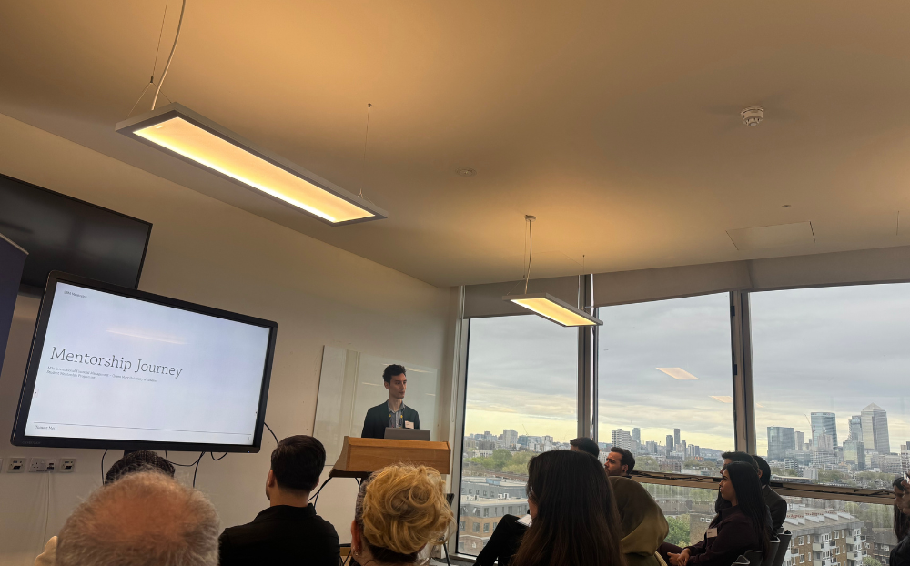 Image of a person speaking infront of a presentation in a room full of people sitting on chairs and the view of the london skyline in the background