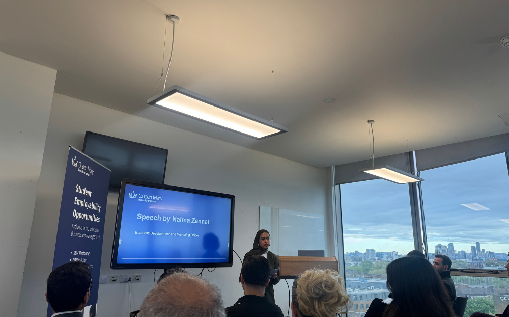 Image of a person speaking infront of a presentation in a room full of people sitting on chairs and the view of the london skyline in the background