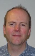 A headshot of Will Hutton. Looking at the camera. Wearing a shirt and with a light grey background.